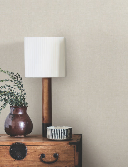 Beige linen-textured wallpaper on the wall of a room; wooden side table with a brown ceramic vase, striped bowl, & tall lamp with a white rectangular shade.