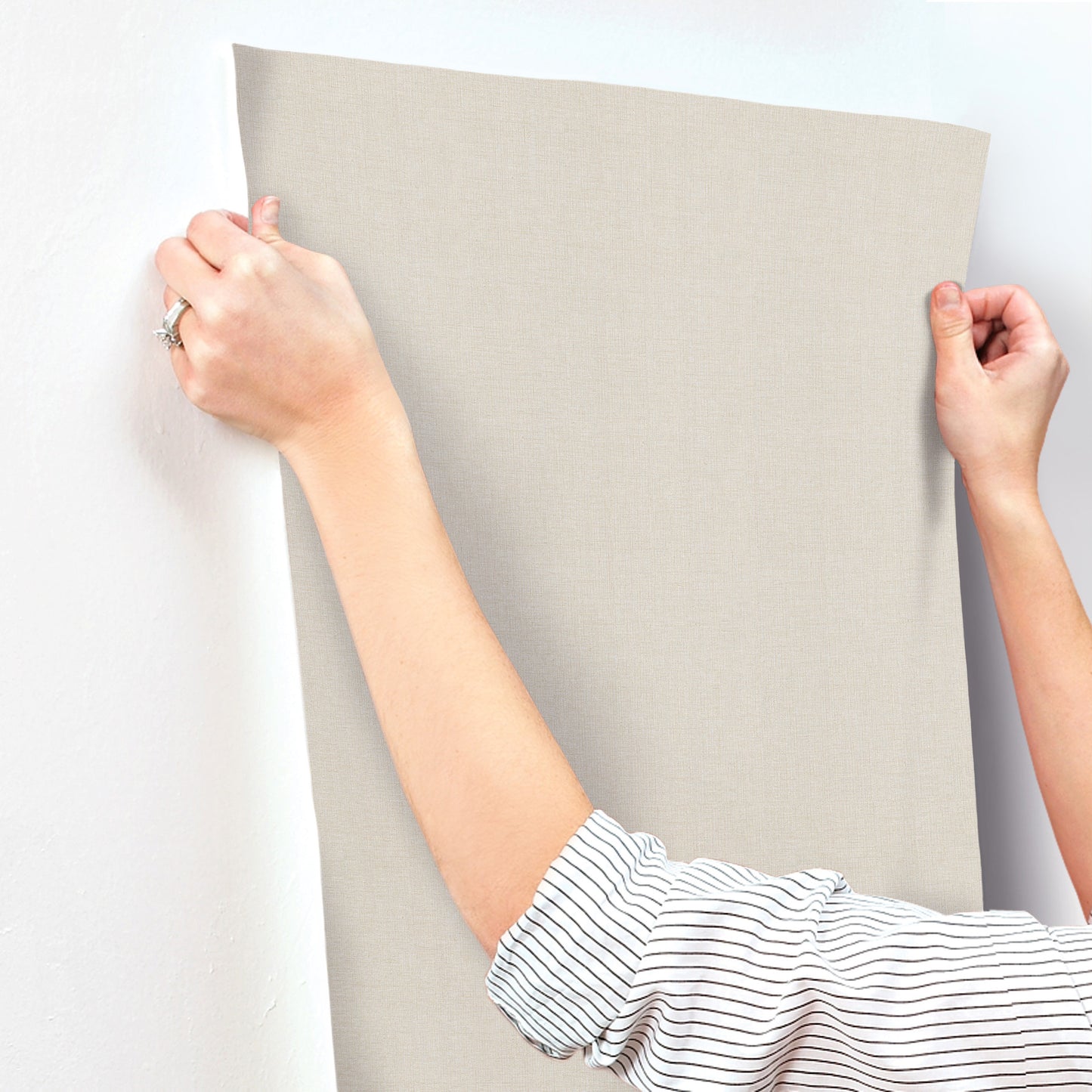 Beige linen-textured wallpaper being stretched taut by two hands; striped shirt sleeves visible.