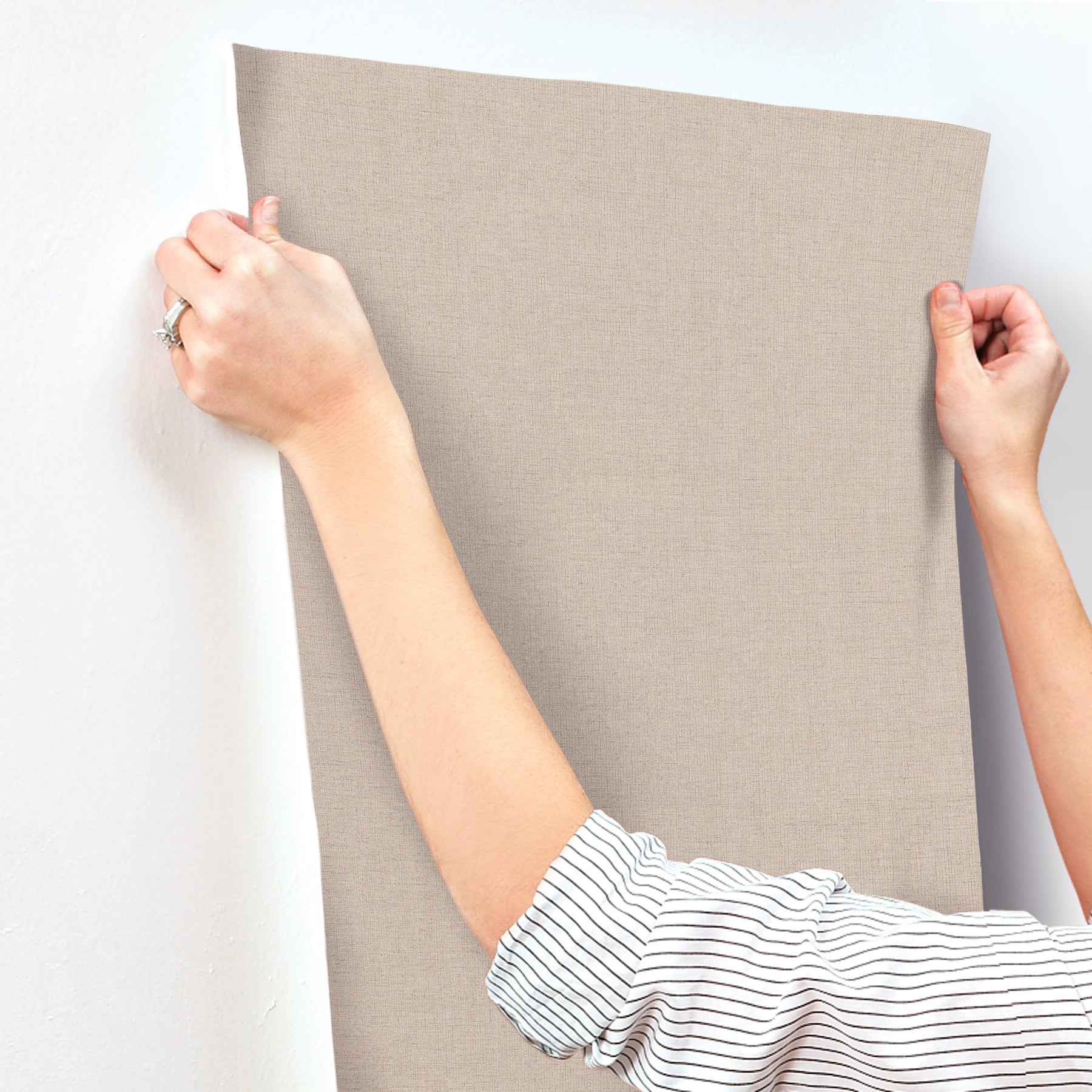 Rectangular beige wallpaper sample with linen-jute texture, held taut by two hands against a white wall.