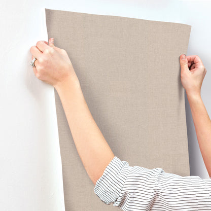 Rectangular beige wallpaper sample with linen-jute texture, held taut by two hands against a white wall.