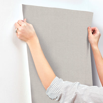 Rectangular, light gray linen-textured wallpaper being held by hands.