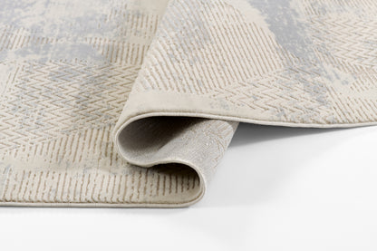 Beige woven area rug with ivory and light gray geometric chevron/diamond pattern, soft low-pile, curled corner for display.