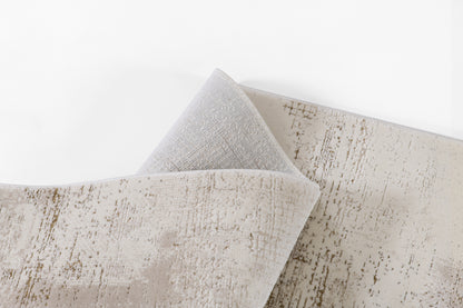 Ivory-beige area rug with subtle distressed brown-taupe pattern, soft plush texture, folded corner reveals gray backing.