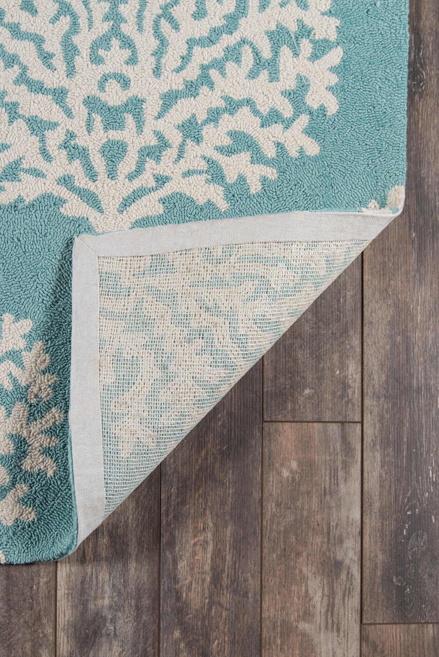 Teal rug with white coral-like pattern, corner folded to reveal pale underside on dark wooden floor.