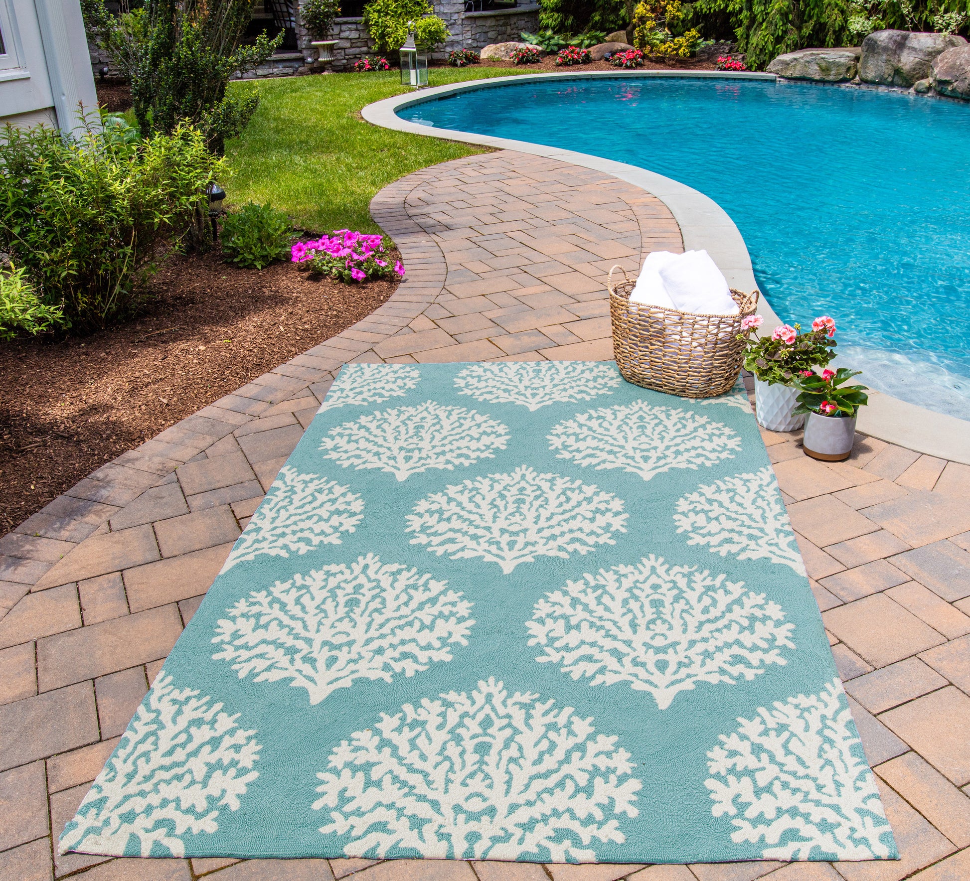 Teal outdoor rug with white tree motif on brick poolside deck; wicker basket with towels nearby by pool.