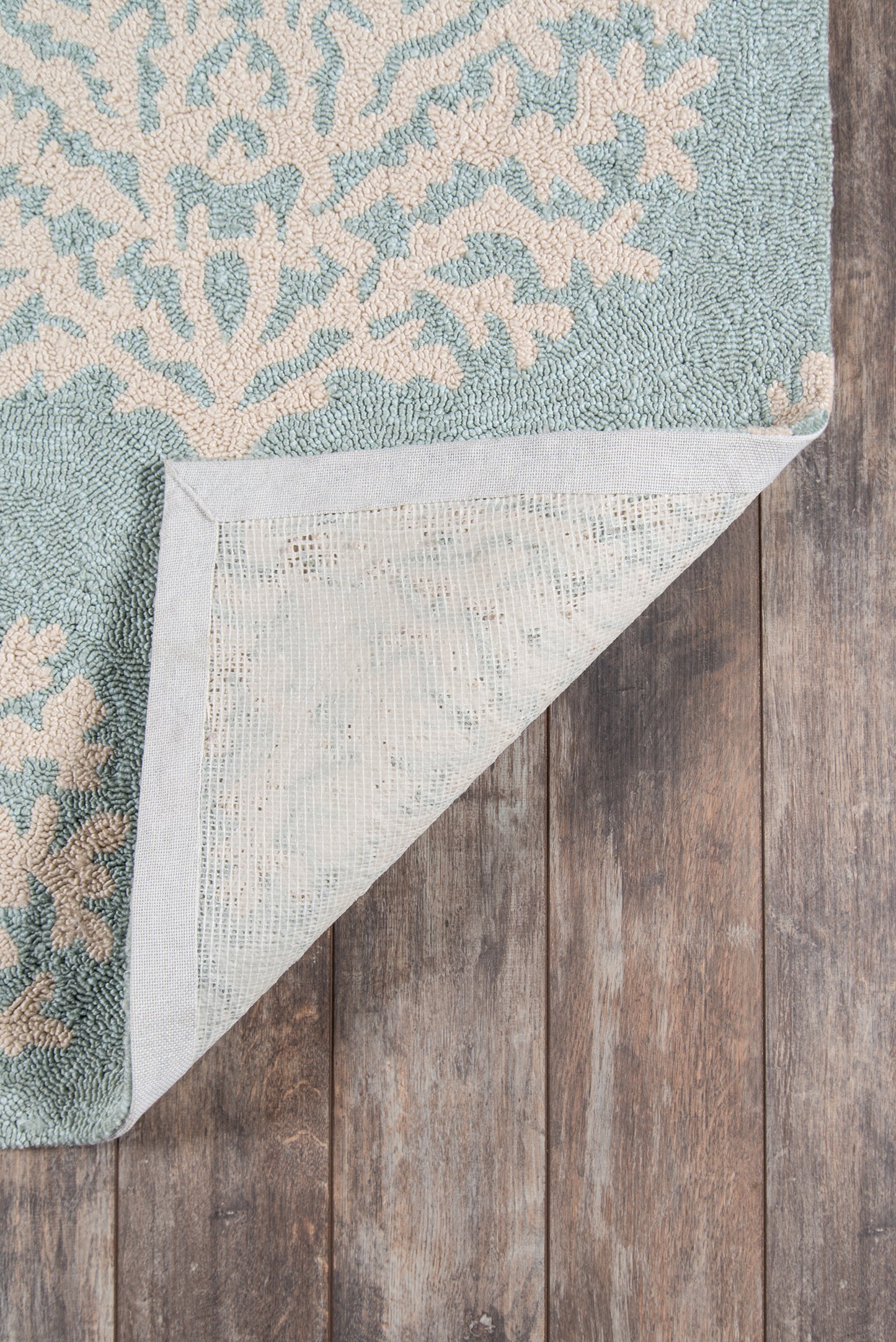 Teal-blue patterned area rug with beige ornamental motif; folded corner shows light backing on wooden floor.
