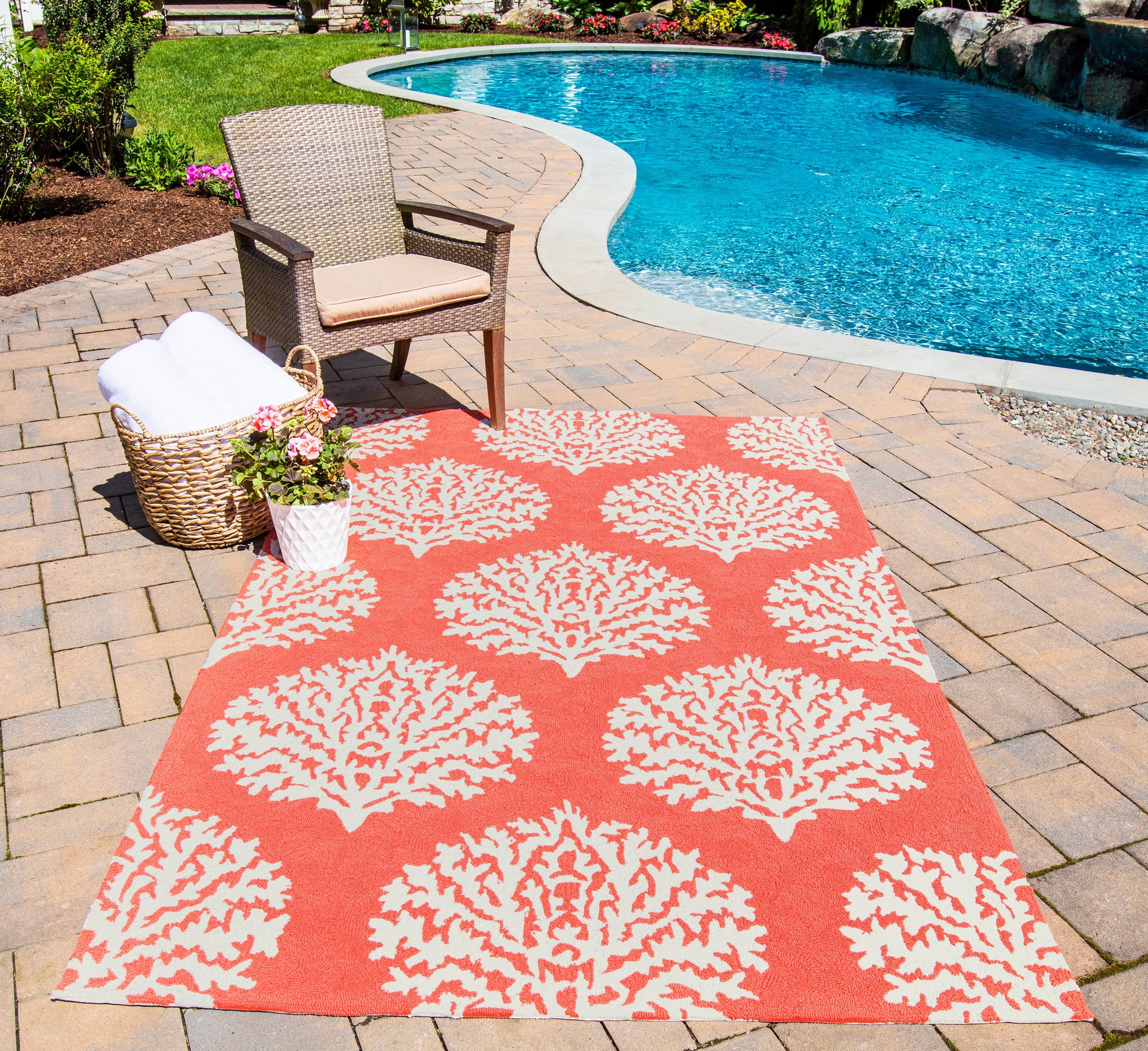 Outdoor poolside setup with an orange rug featuring white coral patterns, a wicker chair with cushion, a towel basket, and potted flowers.