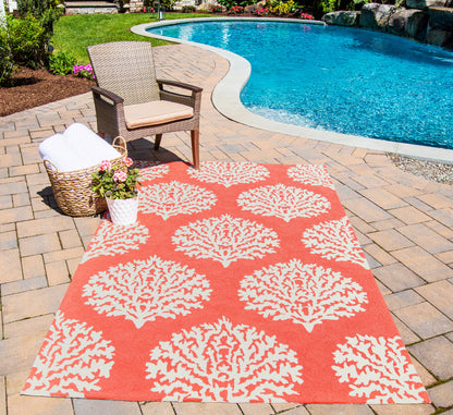 Outdoor poolside setup with an orange rug featuring white coral patterns, a wicker chair with cushion, a towel basket, and potted flowers.