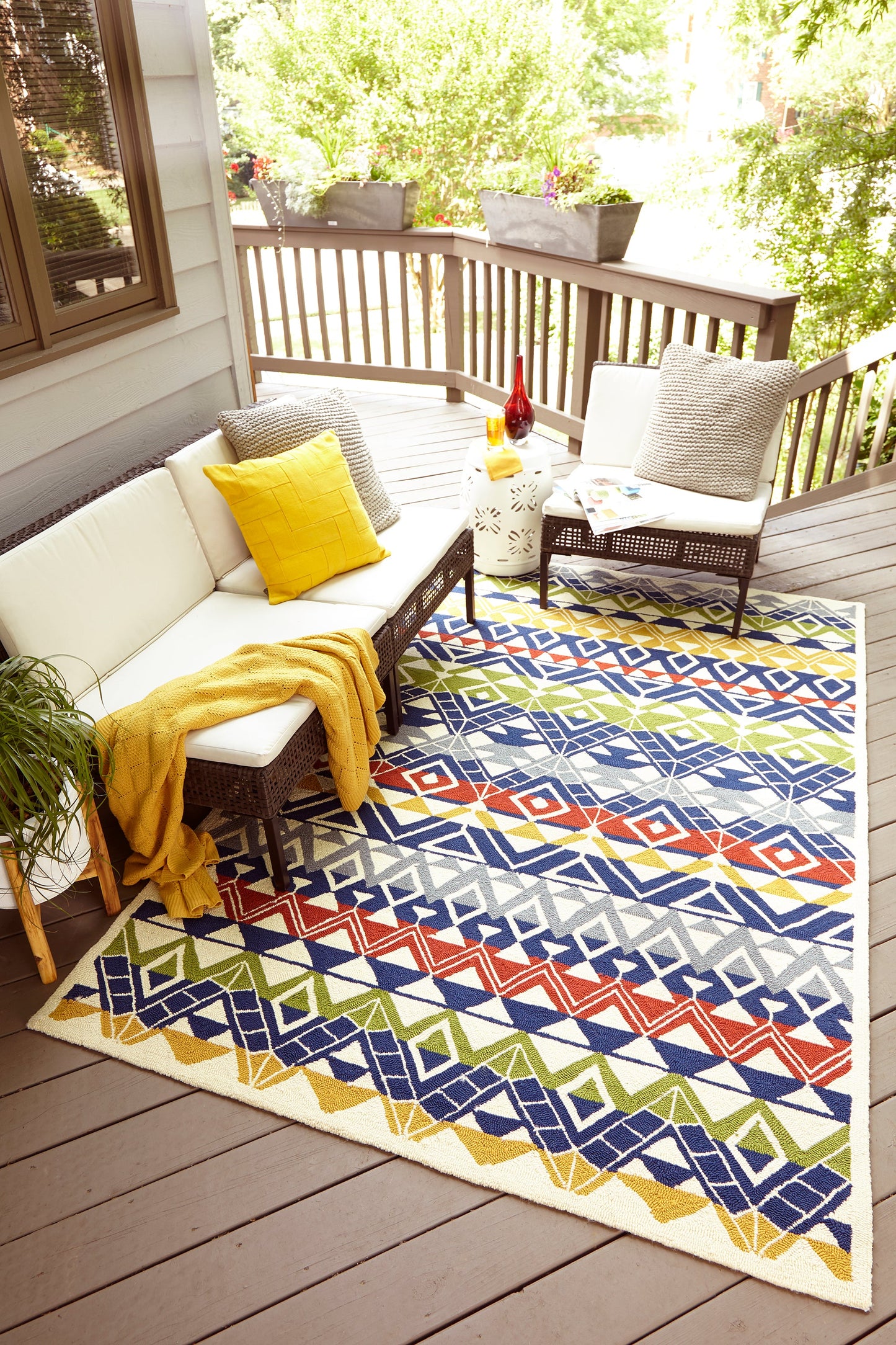 Outdoor geometric chevron rug in blue, orange, green and yellow on a wooden deck, with a white sectional and yellow throw.