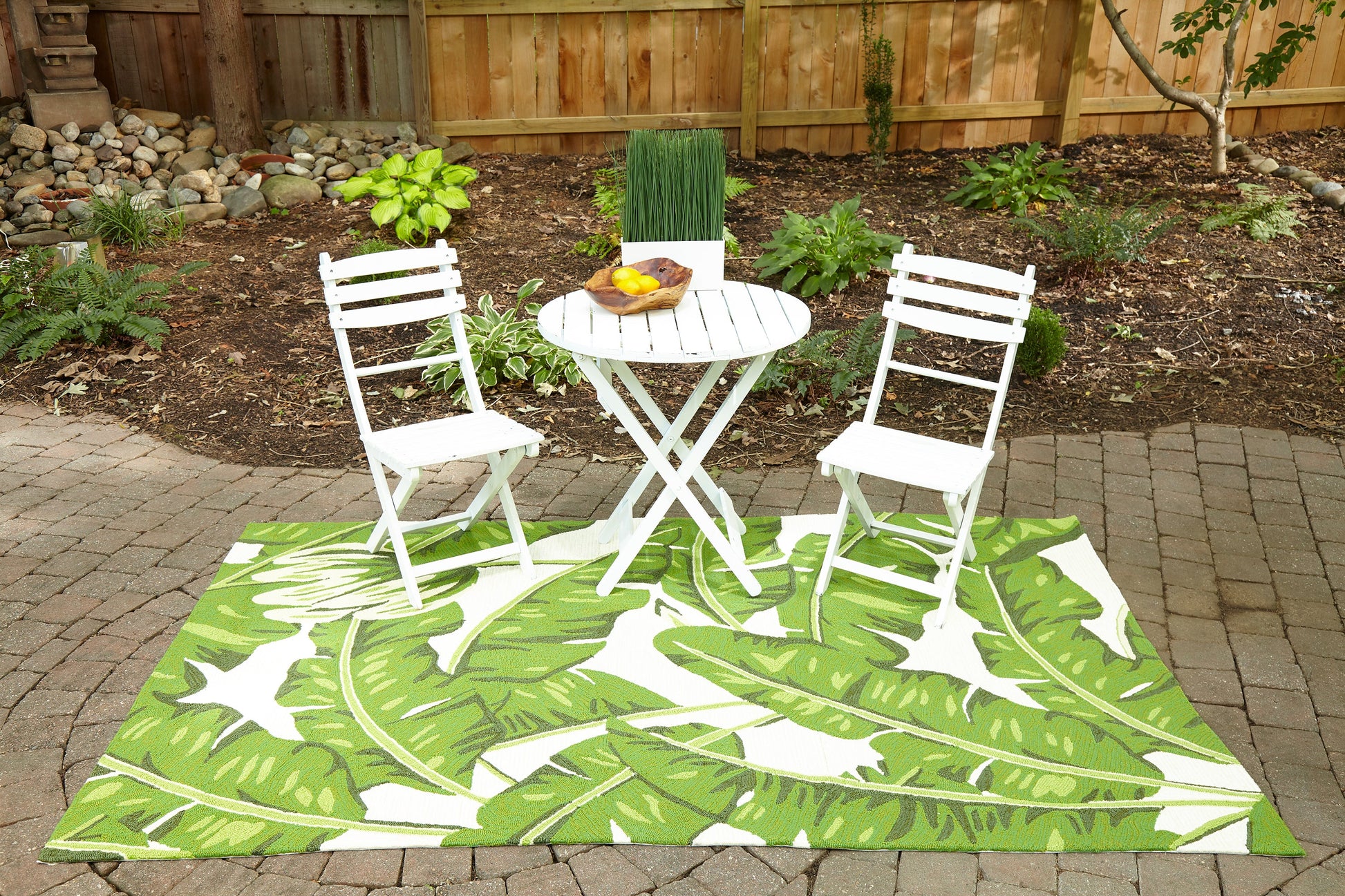 Green leaf-pattern outdoor rug on a garden patio, with a white metal folding bistro table and two chairs.