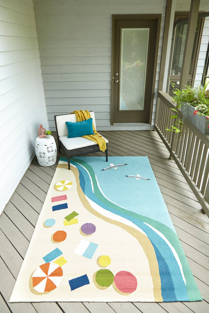 Outdoor balcony rug with beach scene: turquoise waves, sandy shore, colorful shapes; white wicker chair with teal pillow.