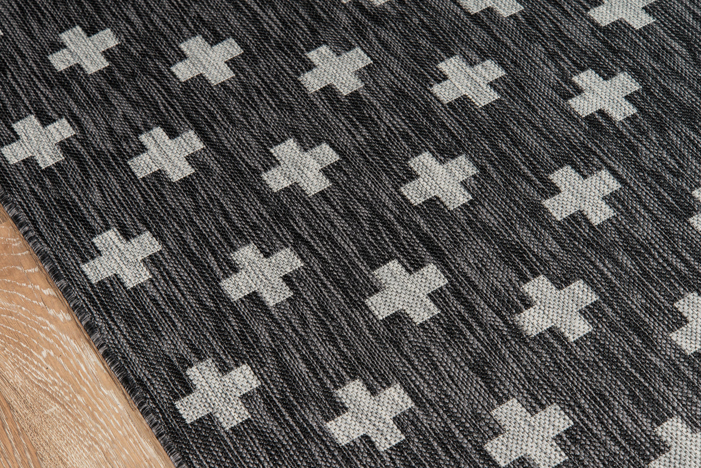 Charcoal woven rug with light gray cross motifs; textured surface and wood floor edge visible.