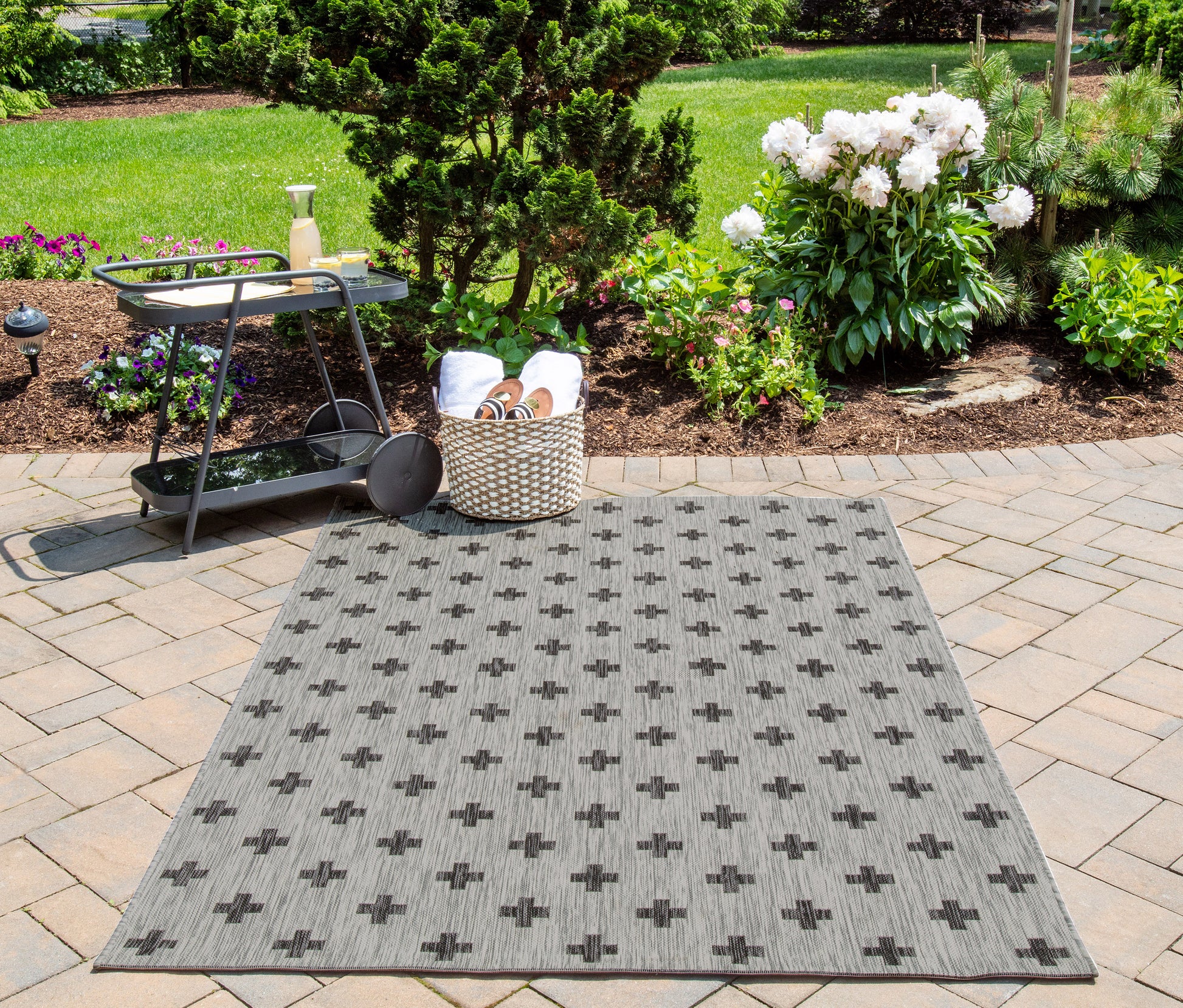 Gray woven outdoor rug with a cross motif on a stone patio, beside a black metal bar cart and lush garden.