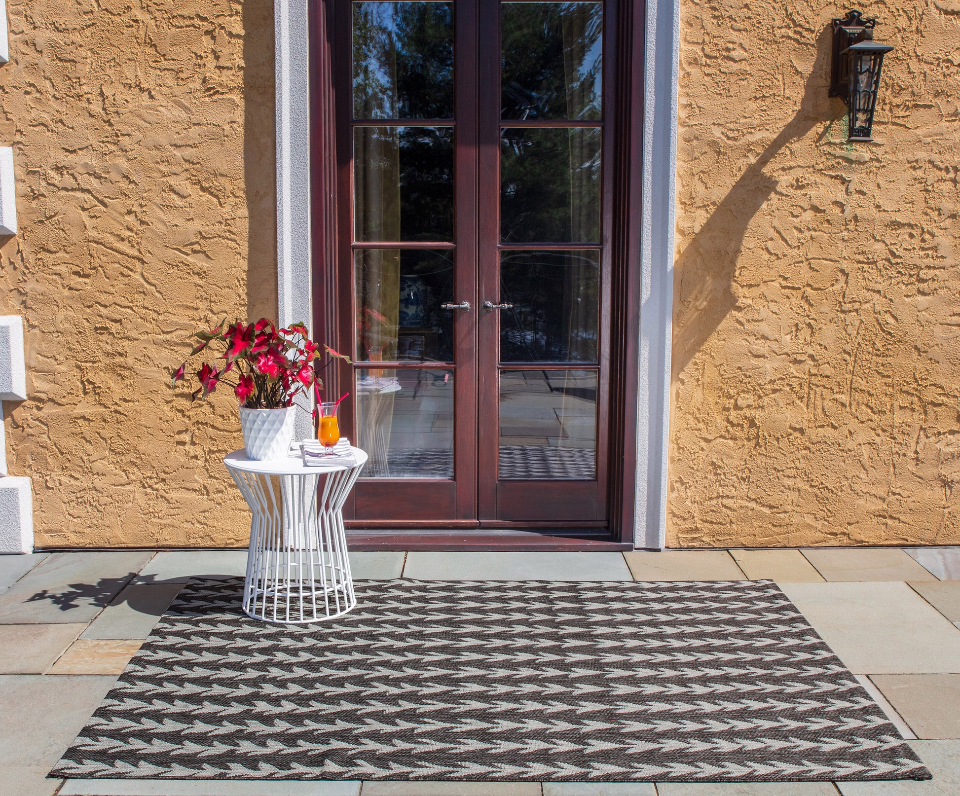 Chevron gray entry mat on tiled porch before orange stucco door; white wire side table with pink flowers and orange vase.
