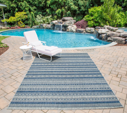 Blue striped outdoor rug on brick patio beside a pool; white lounge chair and side table, lush garden backdrop.