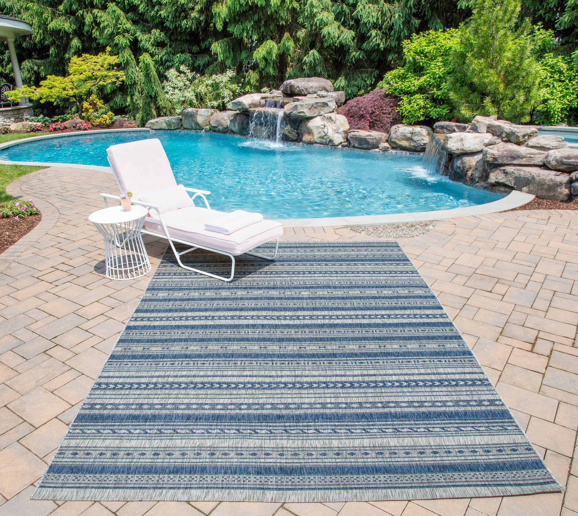 Blue striped outdoor runner rug on a brick pool deck with a white lounge chair and side table beside a waterfall pool (2' x 6').