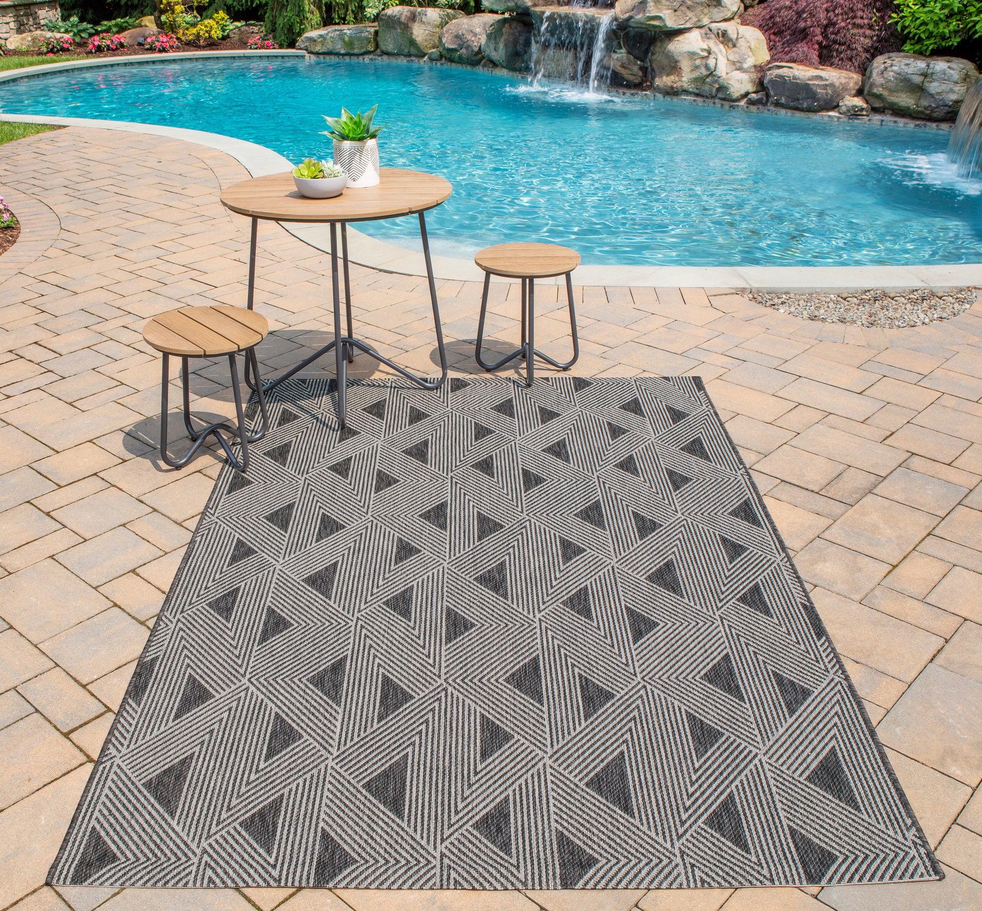 Outdoor poolside patio with geometric gray rug, round wood-top table & two matching stools.