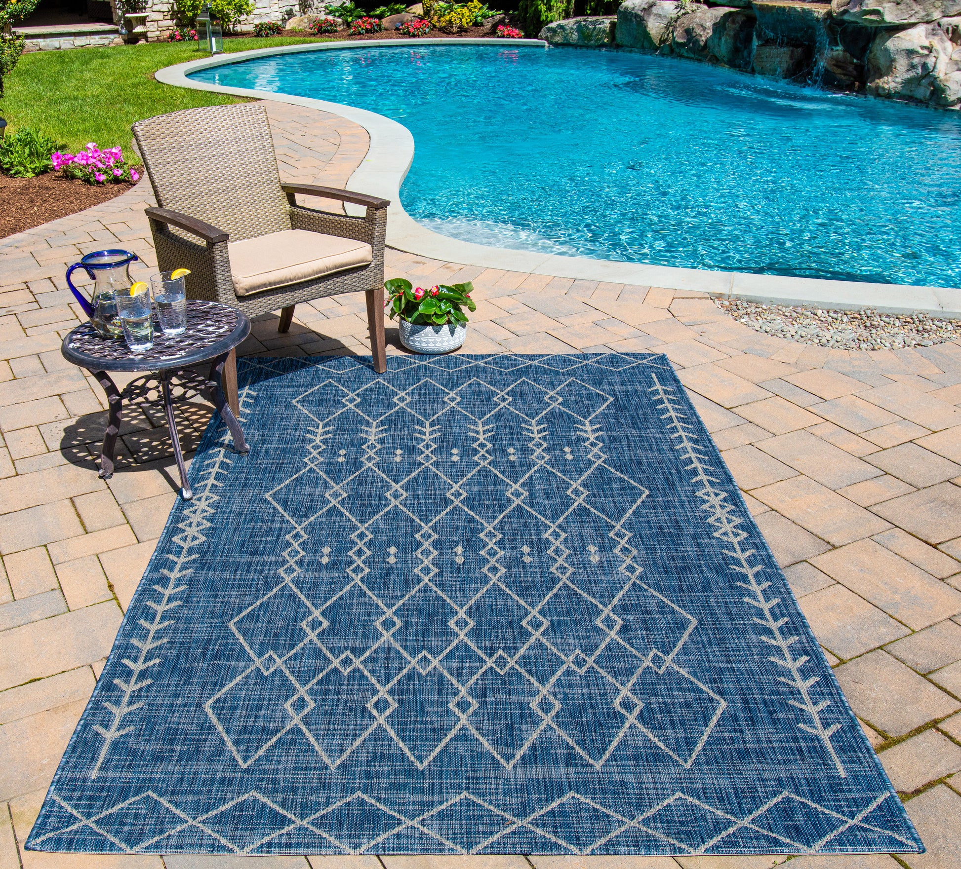Blue geometric outdoor rug on brick poolside patio with wicker chair, small side table, and potted plant.
