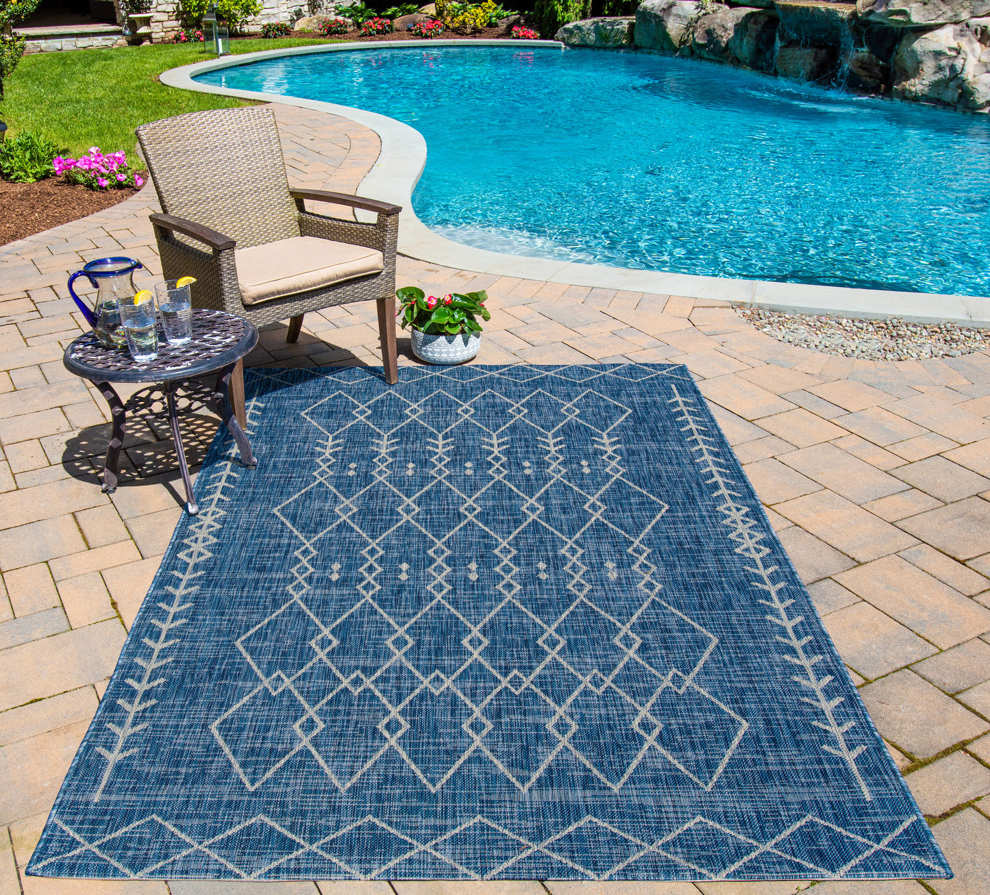 Blue geometric outdoor rug with white lattice pattern on brick patio by pool; wicker chair, round side table, potted plant.