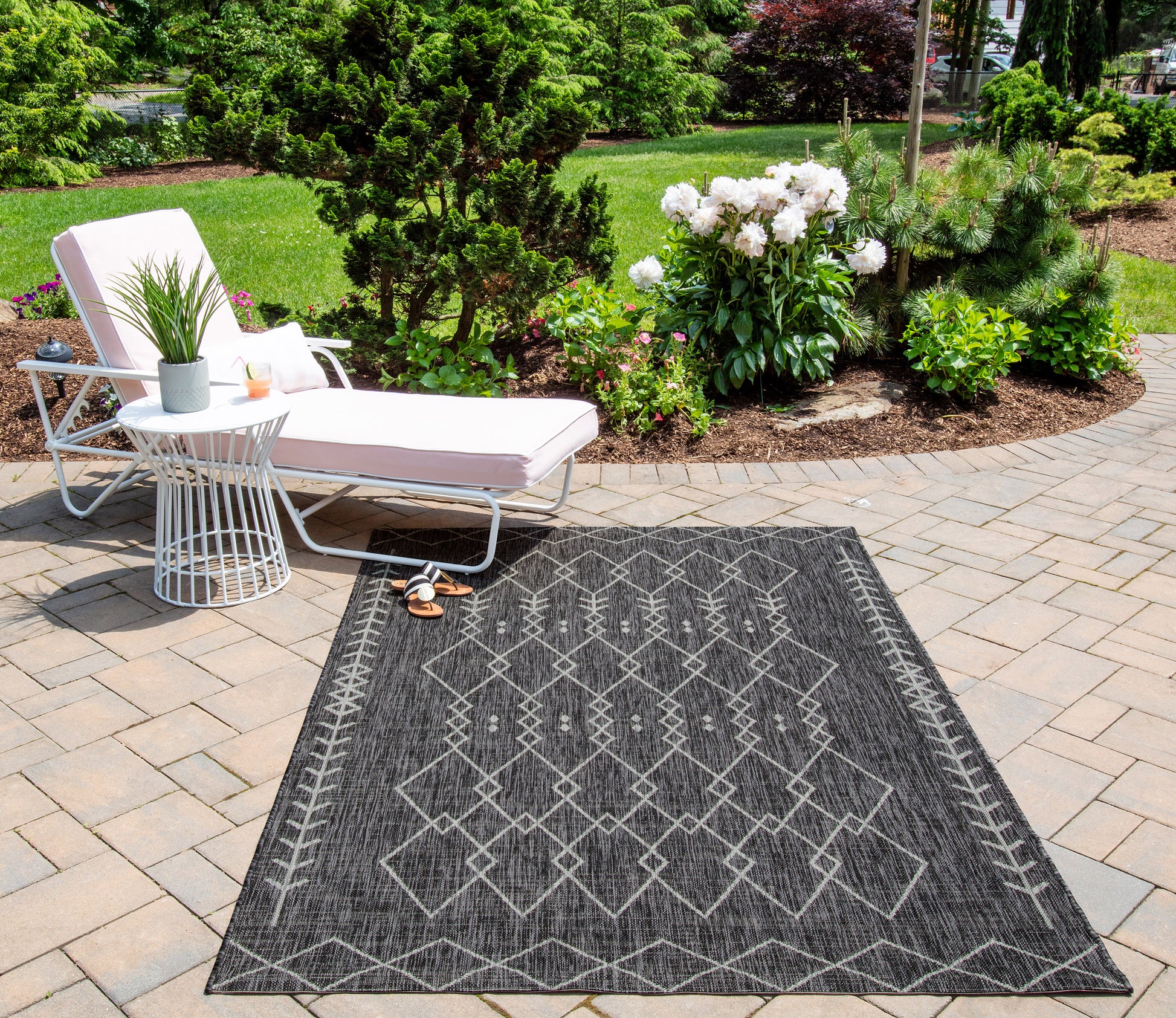 Outdoor dark geometric rug on stone patio beside white chaise lounge with pink cushion, glass side table and greenery.