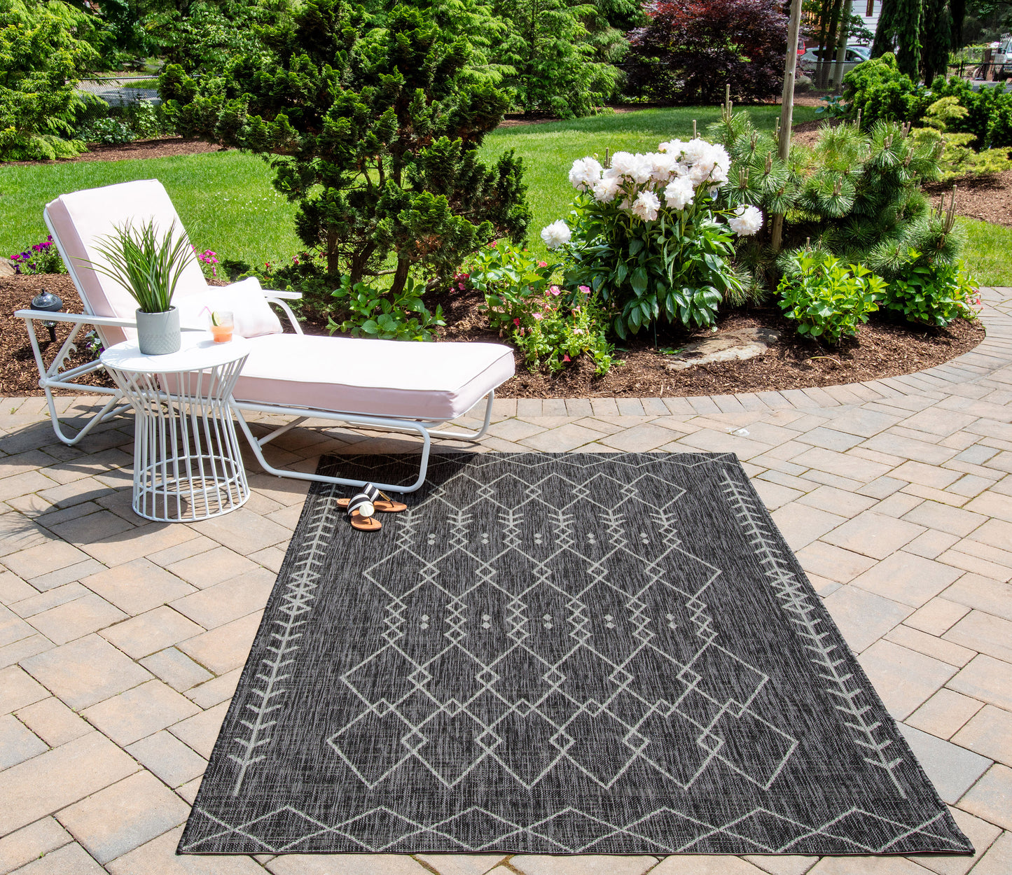 Charcoal geometric outdoor runner rug on a stone patio; white lounge chair, side table, potted plants, and lush garden backdrop.