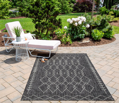 Charcoal geometric outdoor runner rug on a stone patio; white lounge chair, side table, potted plants, and lush garden backdrop.
