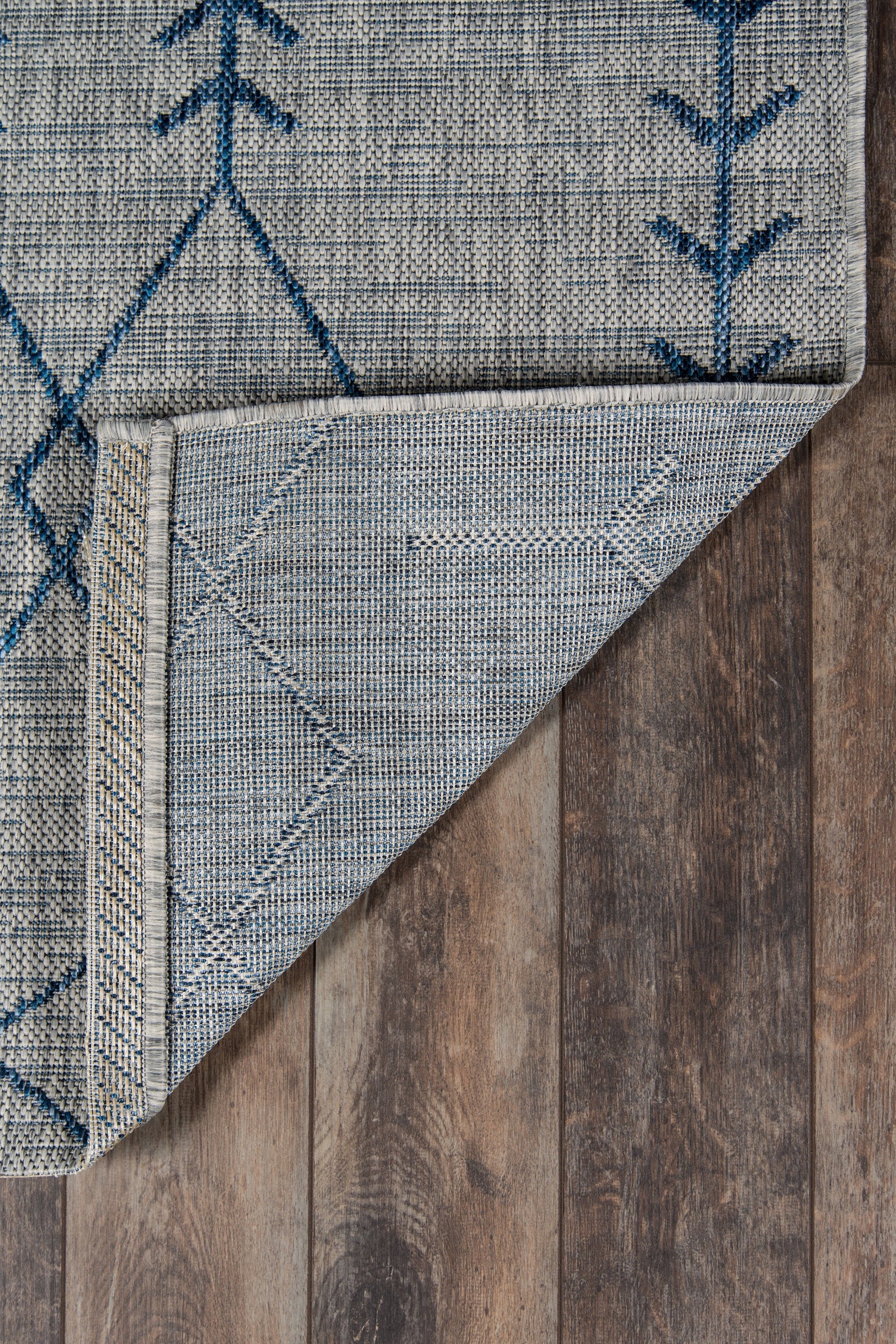 Grey flatweave rug with blue geometric motifs; folded corner shows underside, on dark wood floor.