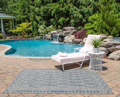 Poolside backyard with white lounge chair, towels, gray geometric outdoor rug, white side table with plant, waterfall rocks & lush greenery.