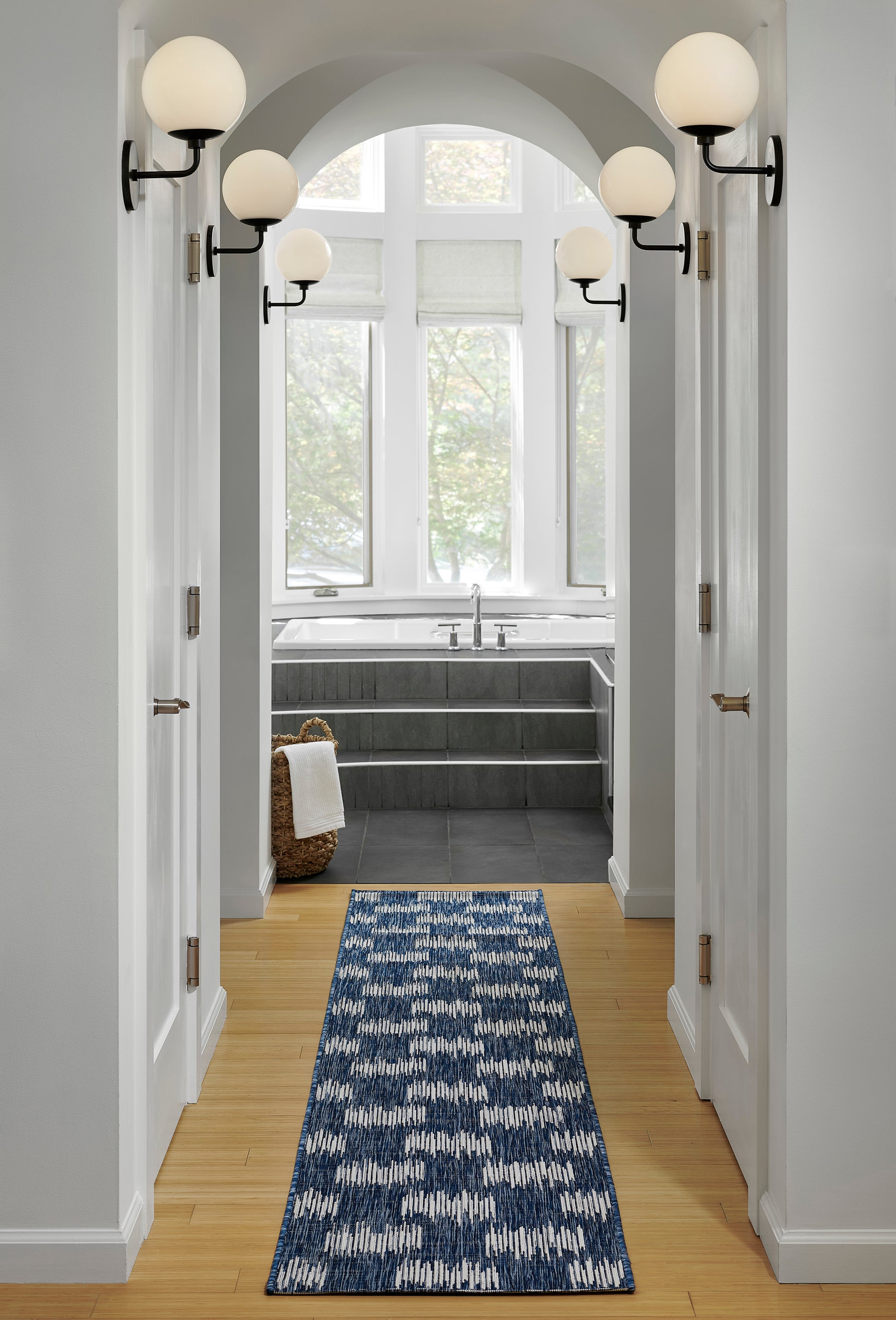 Blue geometric runner rug down a white hallway with an arched doorway, black globe sconces, leading to a grey-tiled bathroom with a tub.