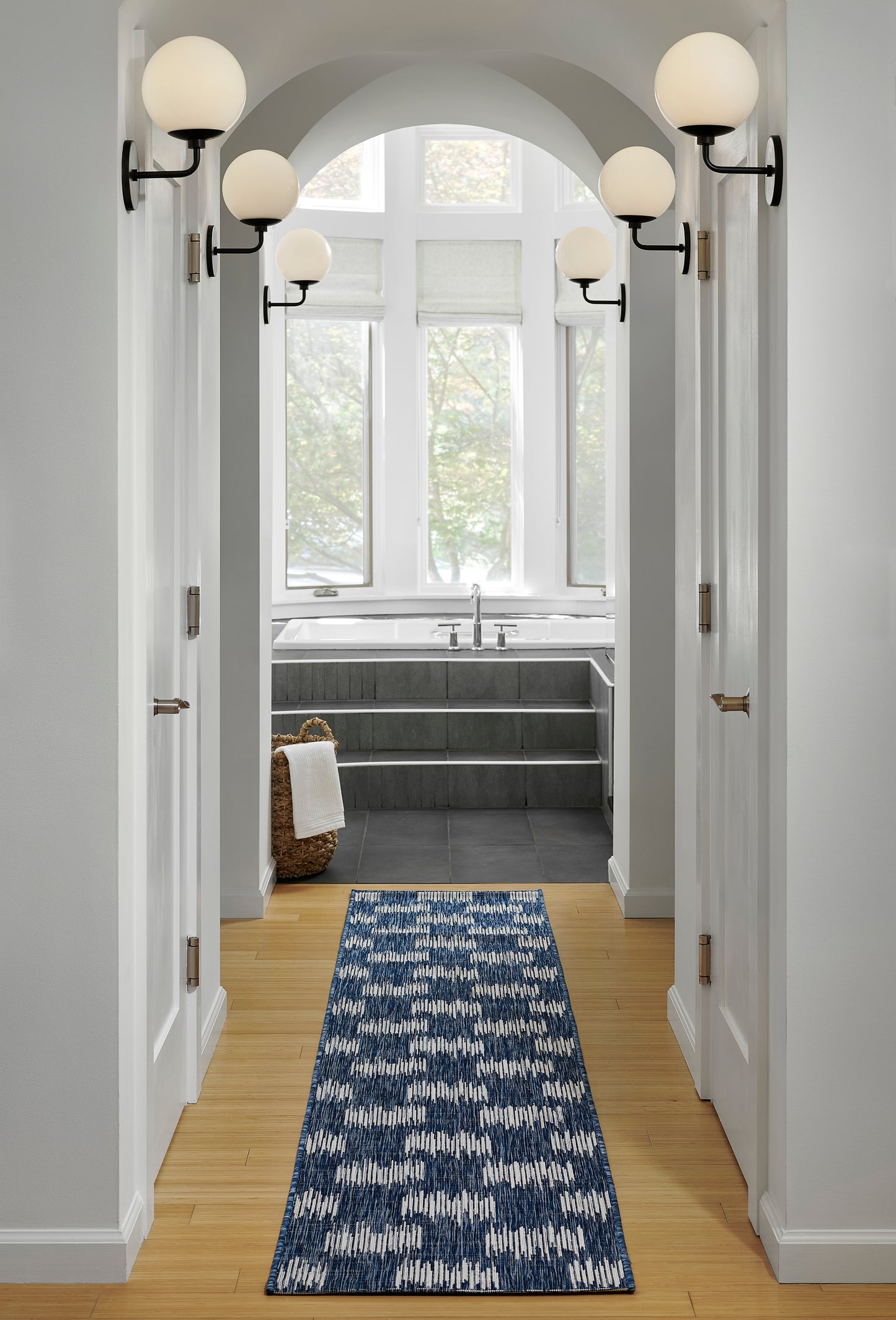 Blue-and-white patterned rug in a white hallway leading to a bath with arched ceiling, globe sconces, and a tub by a window.
