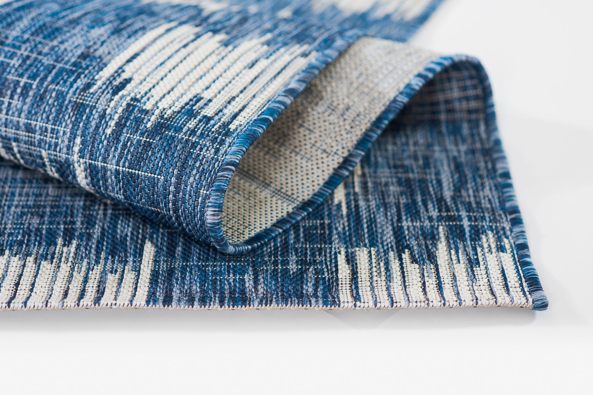 Blue and white woven rug with abstract vertical stripe pattern; folded corner reveals beige underside, modern area rug.