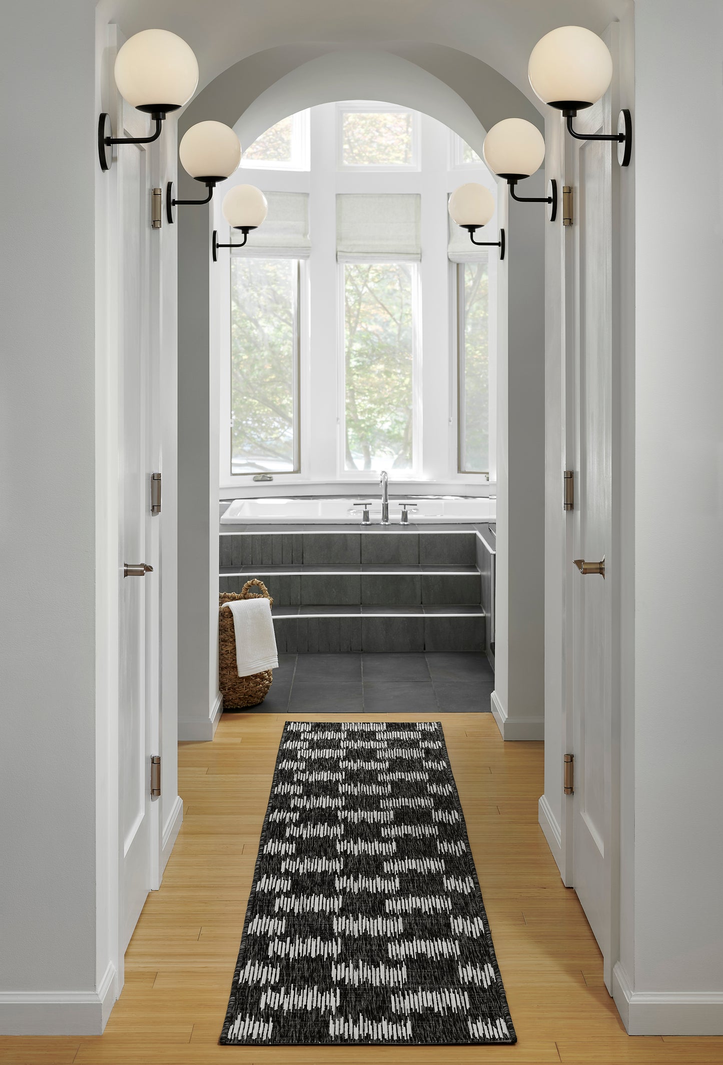 Bright hallway with arched openings, black metal globe sconces, light wood floor, black-white geometric runner, leading to a bathroom.