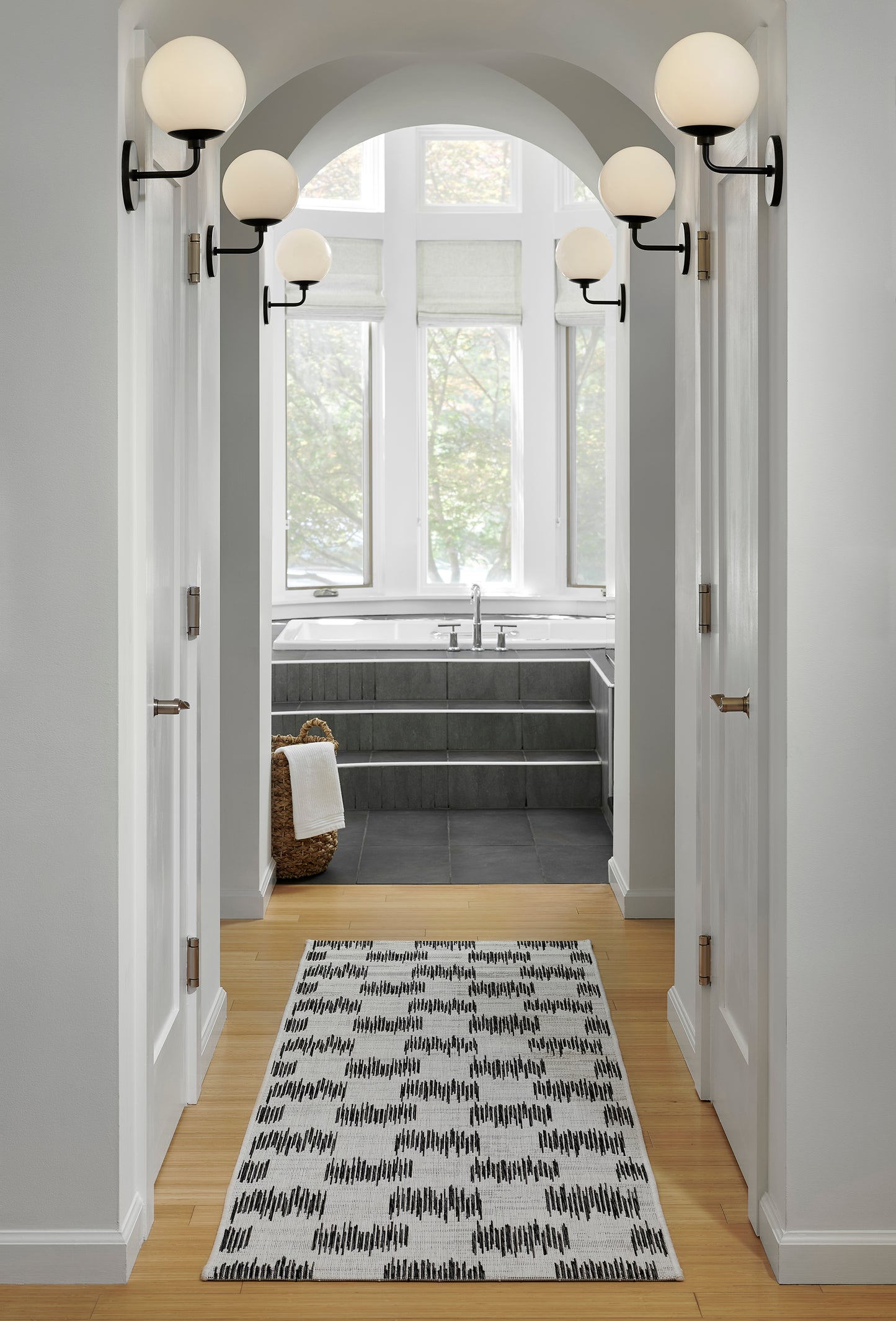 Cream rug with black geometric pattern in a light-gray hallway toward a gray-tiled bath with globe sconces and arched ceiling.