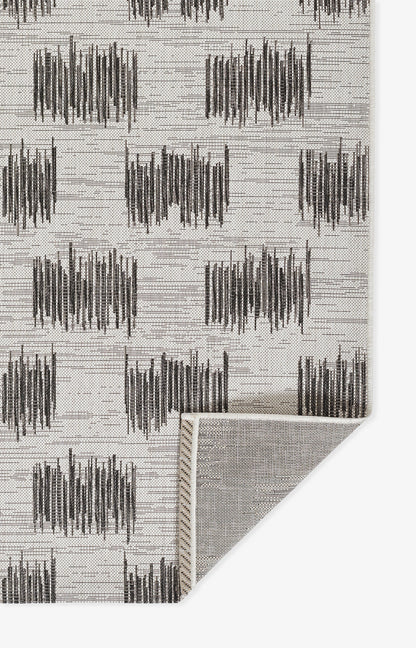 Beige rug with repeating dark charcoal brushstroke blocks; soft woven texture, corner folded to show light underside.
