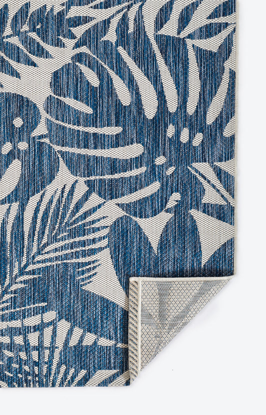 Blue woven rug with white tropical leaf pattern; textured fabric, folded corner shows beige backing.