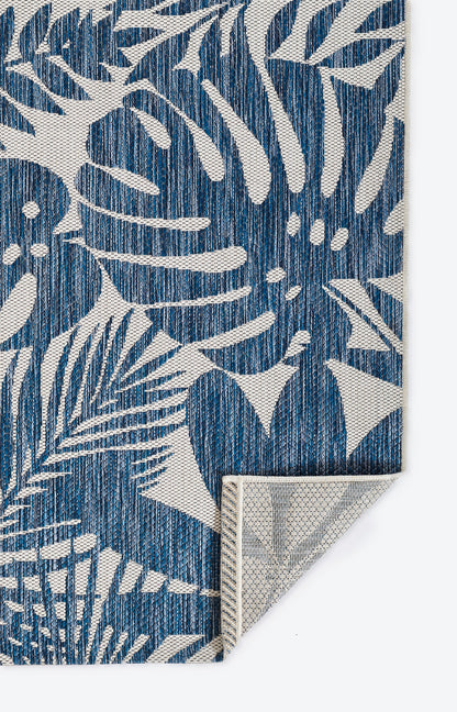 Blue rug with white tropical leaf pattern; woven texture & folded corner showing light underside.