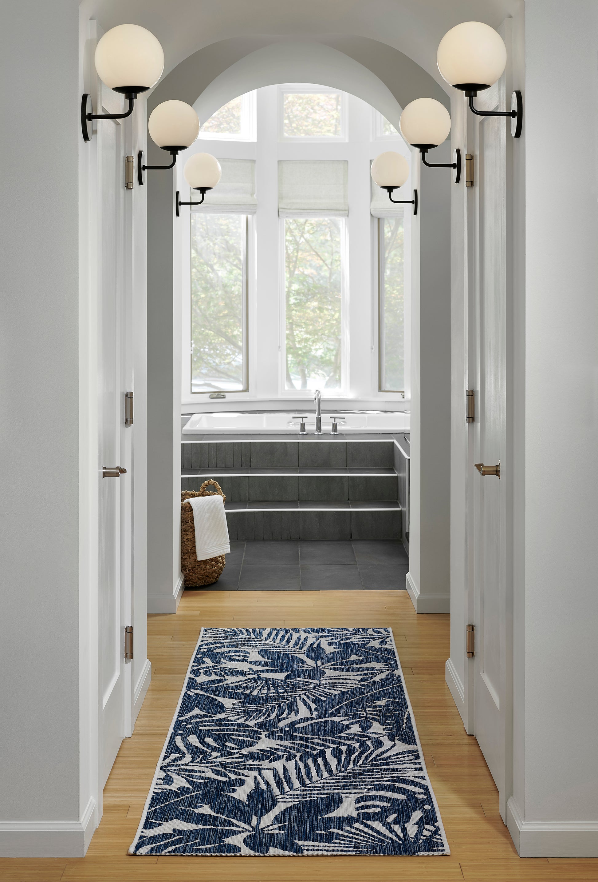 Navy blue palm-leaf print rug in a light wood hallway leading to a grey-tiled bath with arched window and globe wall sconces.