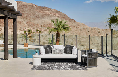 Outdoor lounge with white cushioned sofa, black/white pillows, gray leaf rug, glass railing, pool and desert hills.