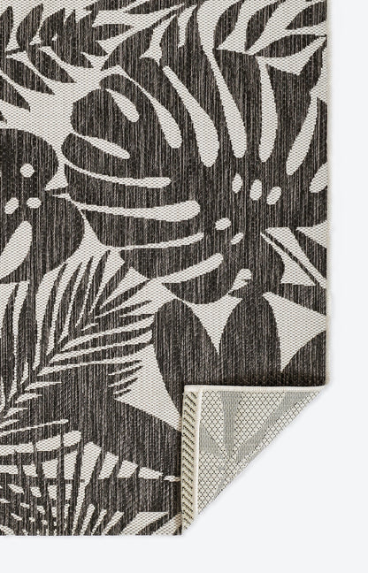 Charcoal and cream botanical leaf rug; woven texture with monstera/palm leaves, folded corner.