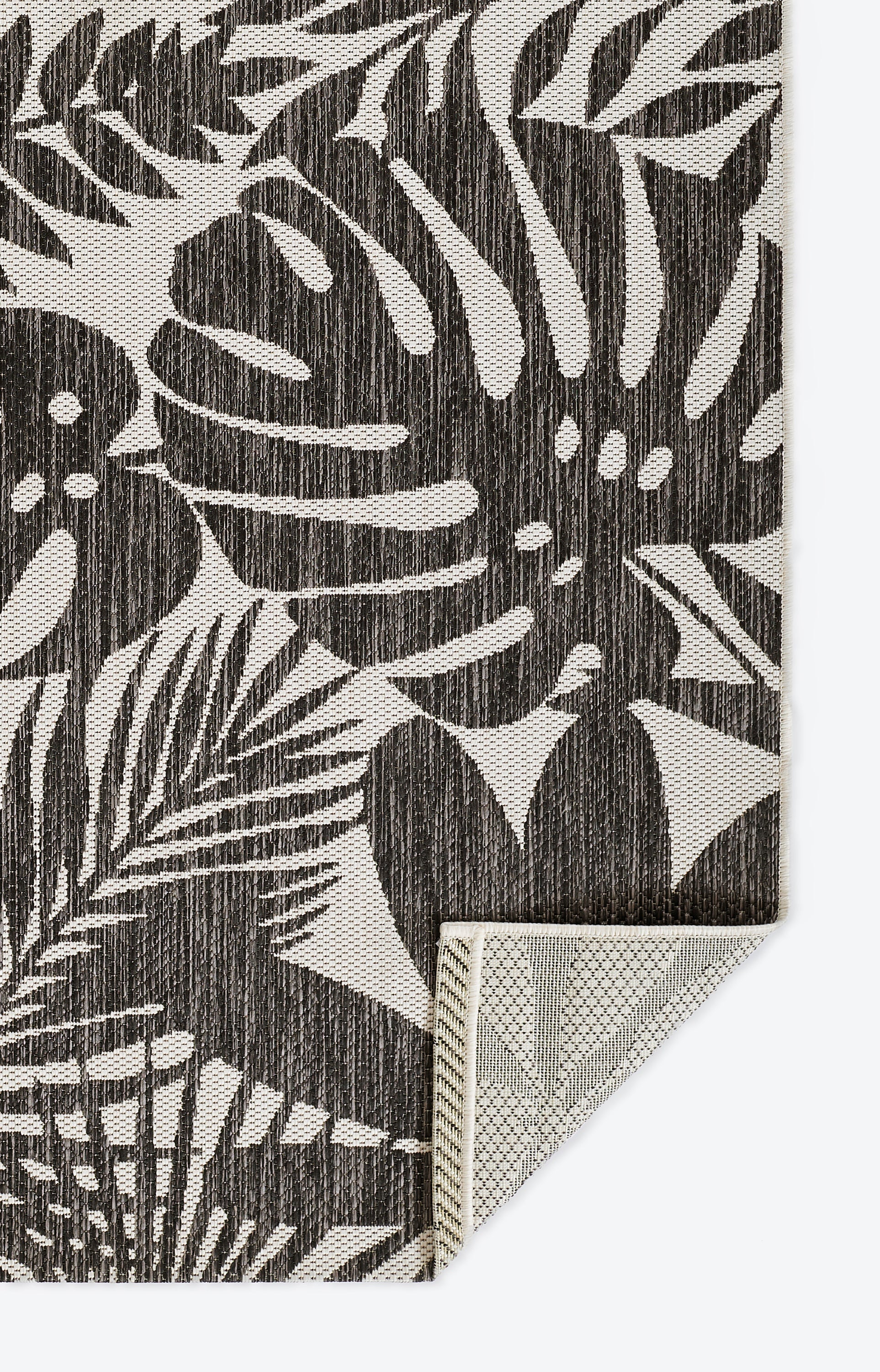 Charcoal botanical leaf rug with monstera and palm fronds on a beige backdrop; textured textile with a folded corner.