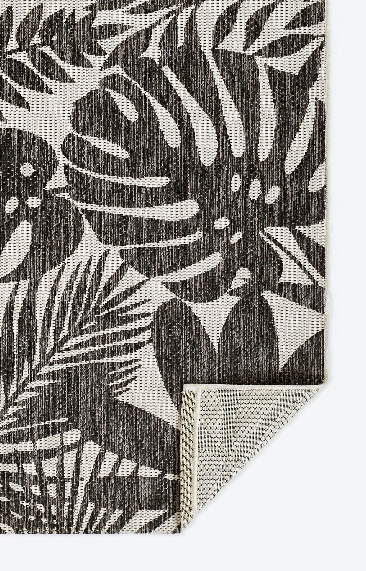 Charcoal palm-leaf patterned rug on beige base; woven textile with textured fibers, corner folded to reveal underside.