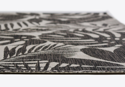 Close-up of a flat-woven rug with black background and white tropical leaf pattern, textured fiber edge visible.