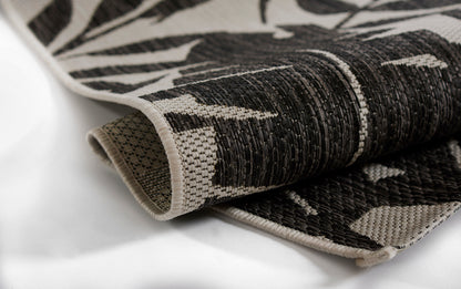 Rolled black and ivory woven rug with botanical leaf pattern; textured fabric and beige bound edge.