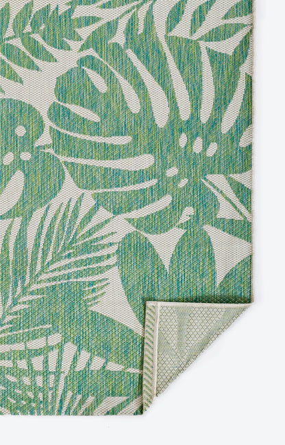 Rectangular woven rug with green tropical monstera leaves on beige; folded corner shows light backing.