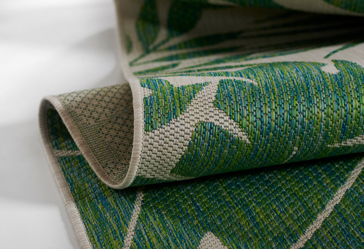 Woven area rug in green and teal with beige background; textured fabric, leaf-pattern design, folded edge.