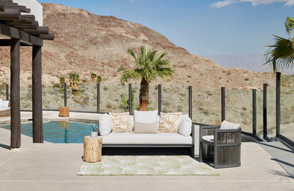 Green palm-leaf rug on a poolside patio with a white sofa, dark wicker chairs, palm trees, glass railing and beige tiles.