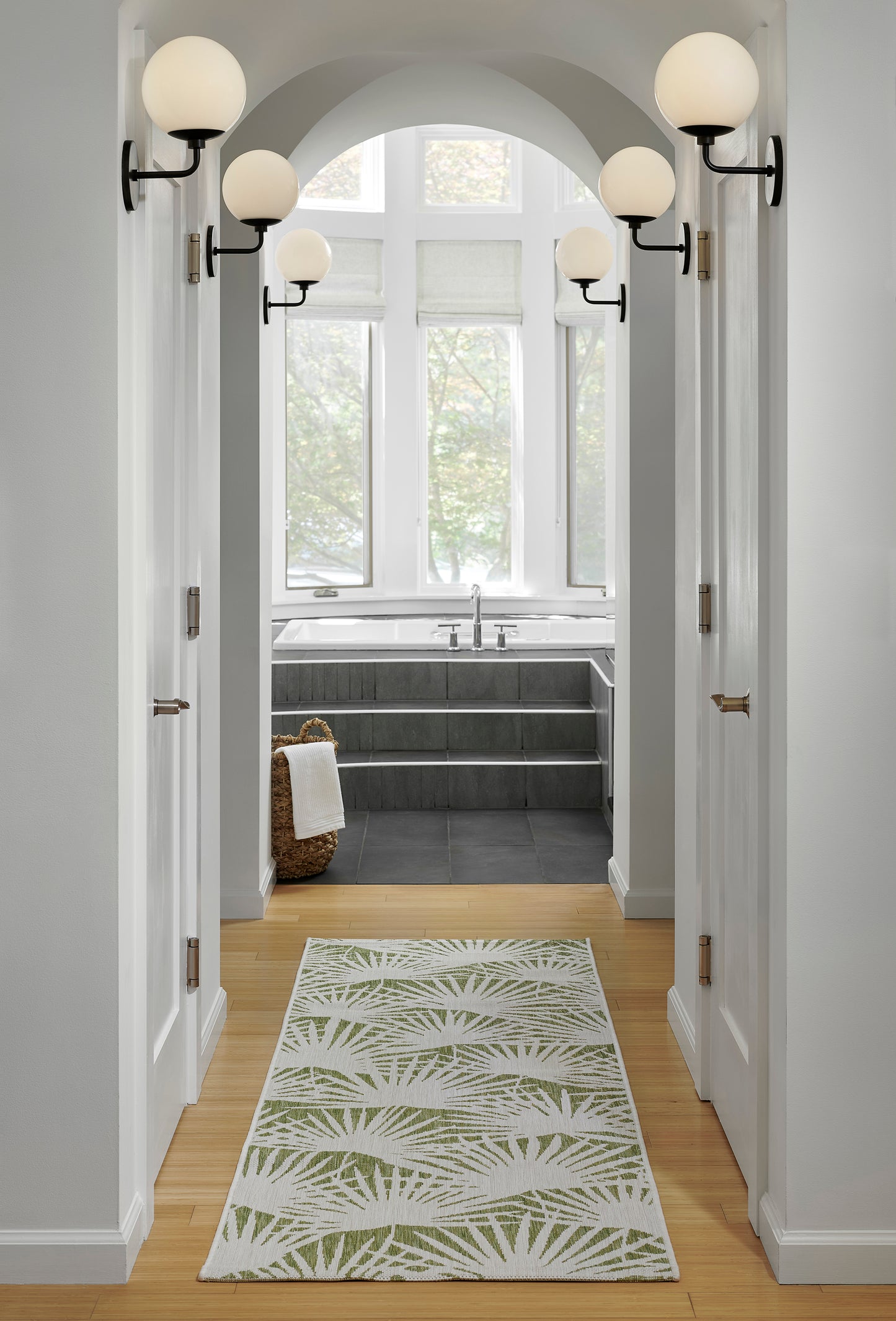 Hallway to bathroom with arched doorway, white walls, light wood floor, green palm-leaf rug, globe sconces on each side.