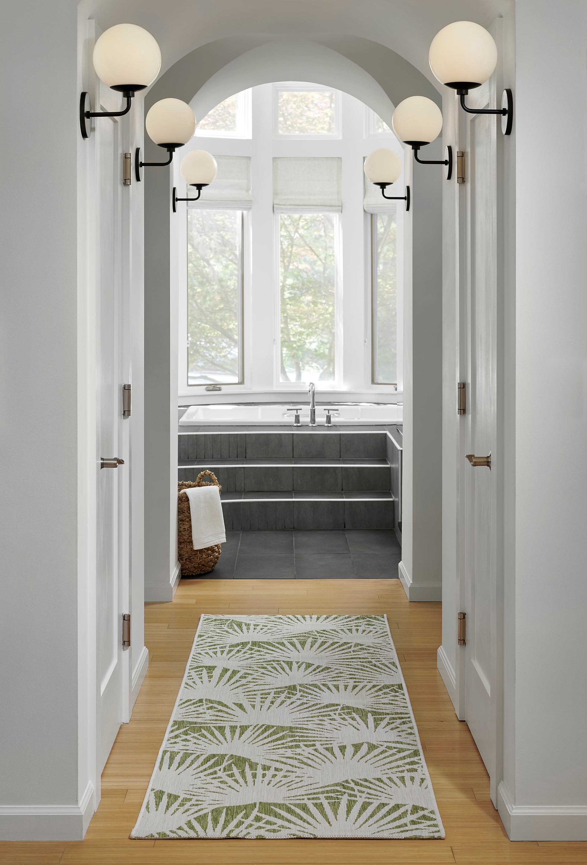 Green palm-leaf rug, 3' x 5', in a light wood hallway with an arched doorway and brass wall sconces.