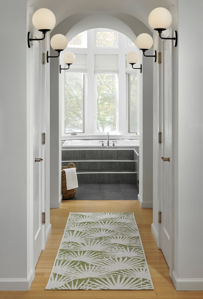 White hallway leads to a bathroom; arched doorway, globe wall sconces, light wood floor, green palm-leaf runner rug.