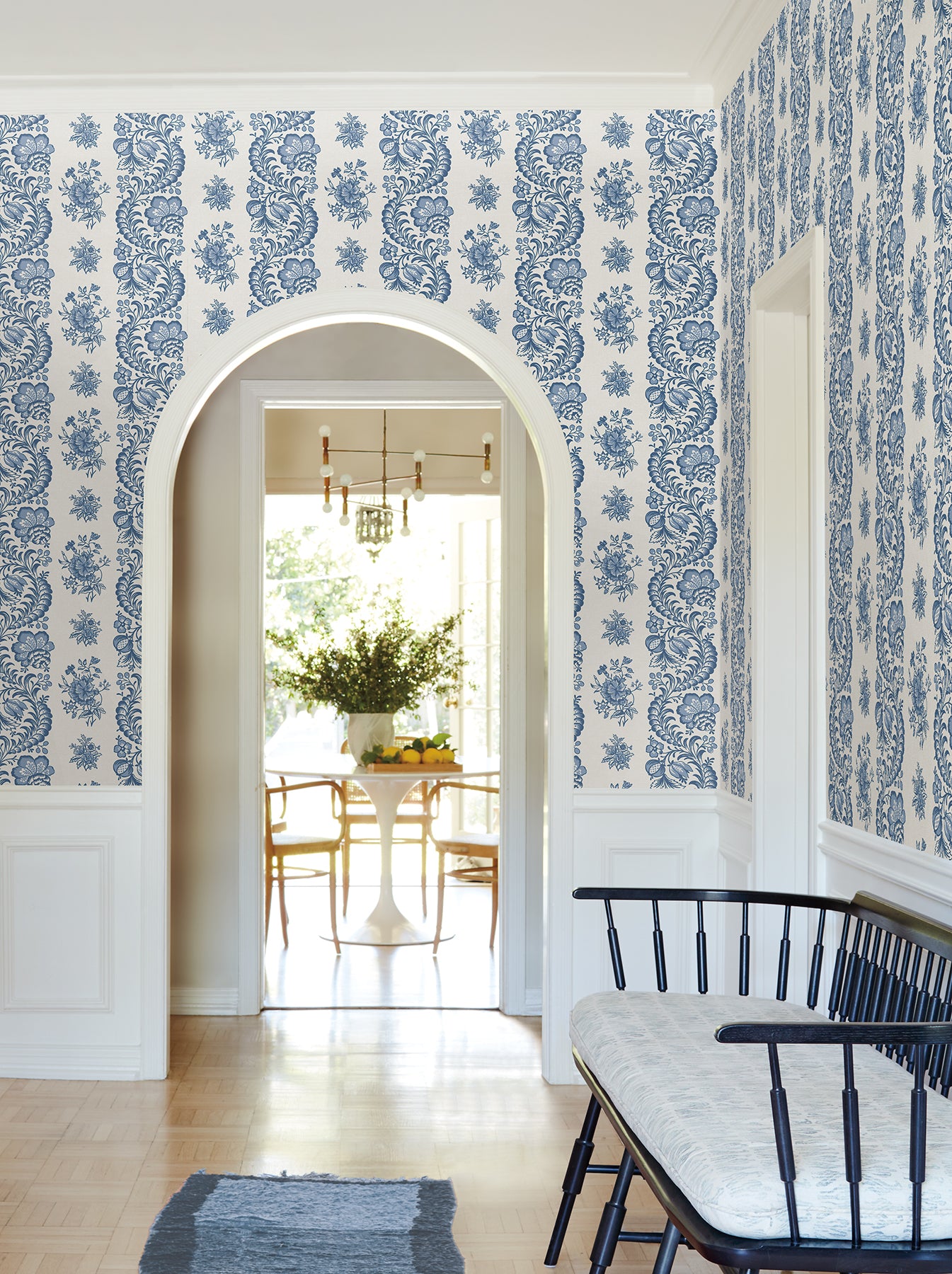 Blue and white damask wallpaper in a white wainscoted foyer; arch doorway to a sunlit dining room with pedestal table.